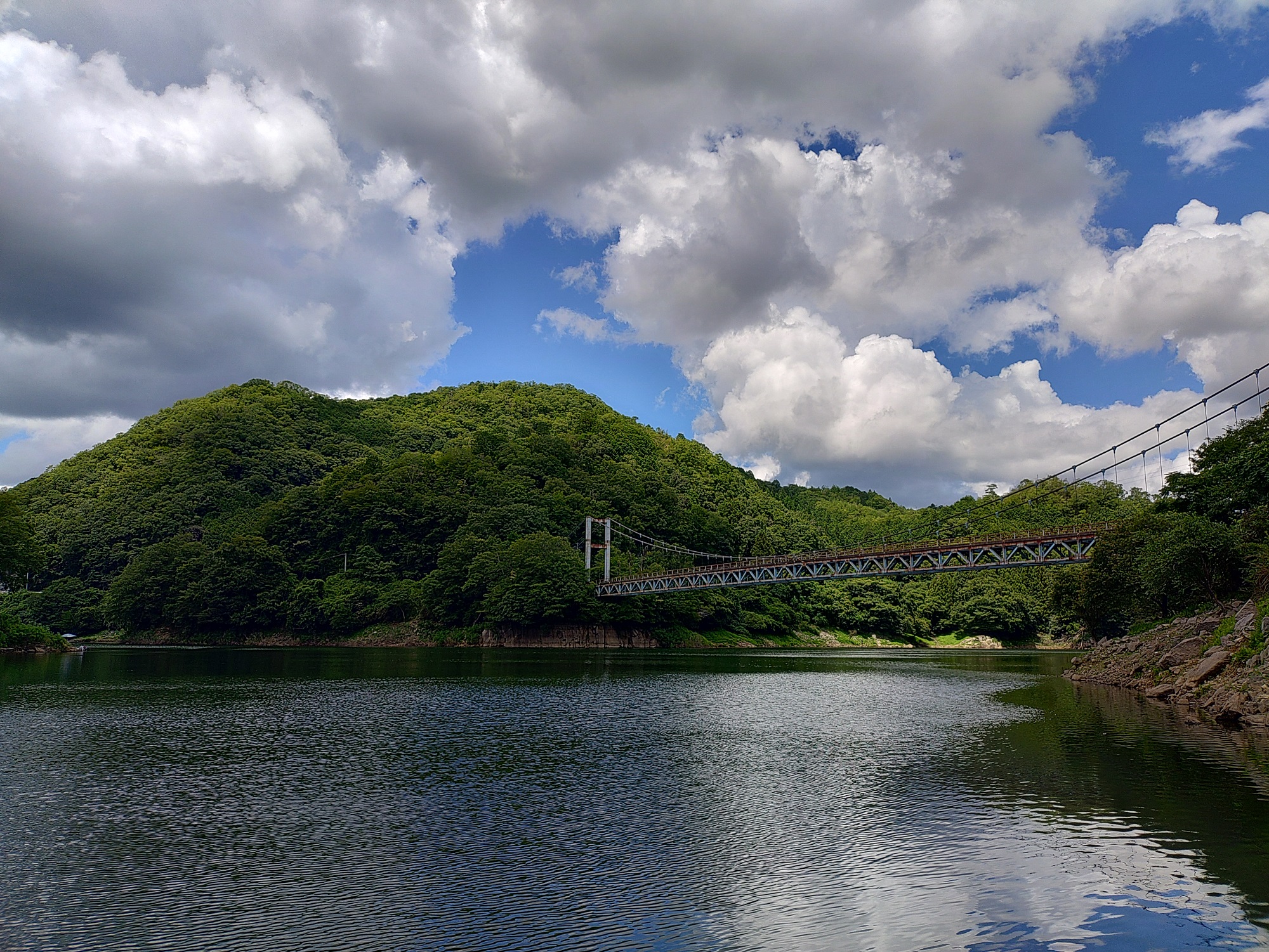豊かな緑に囲まれた湖面に吊り橋が架かり、青空と大きな雲が広がる水辺の風景